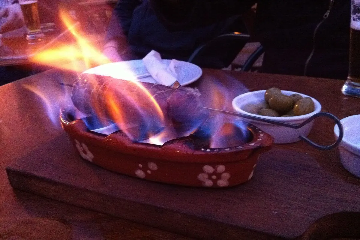 A chouriço assado, a smoked sausage flaming in a red-patterned clay dish on a wooden board, with olives and a glass beside it