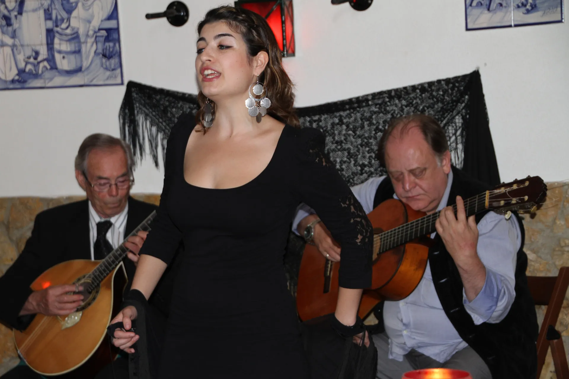 A fado singer in a black dress between two guitarists, one holding a pear-shaped Portuguese guitar and the other a six-string guitar, in a small Lisbon fado house with blue tiles and a red lantern on the wall