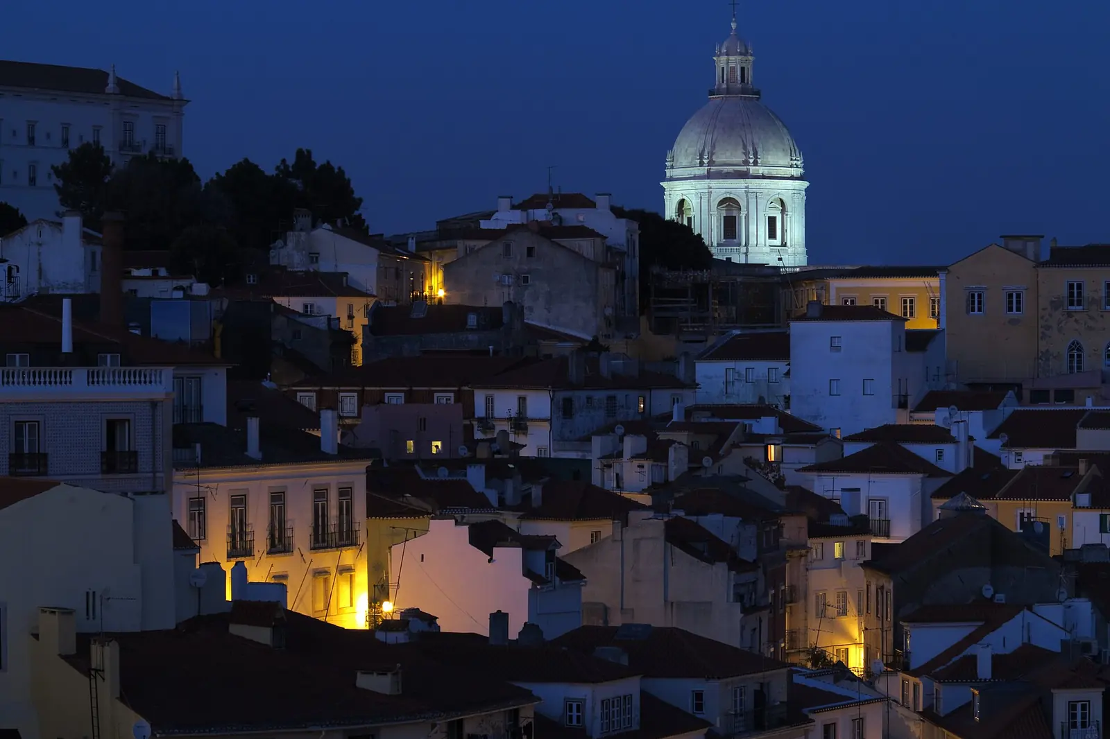 Whitewashed and terracotta-roofed houses on the slope of Alfama at dusk, with the dome of the National Pantheon lit against a deep blue sky