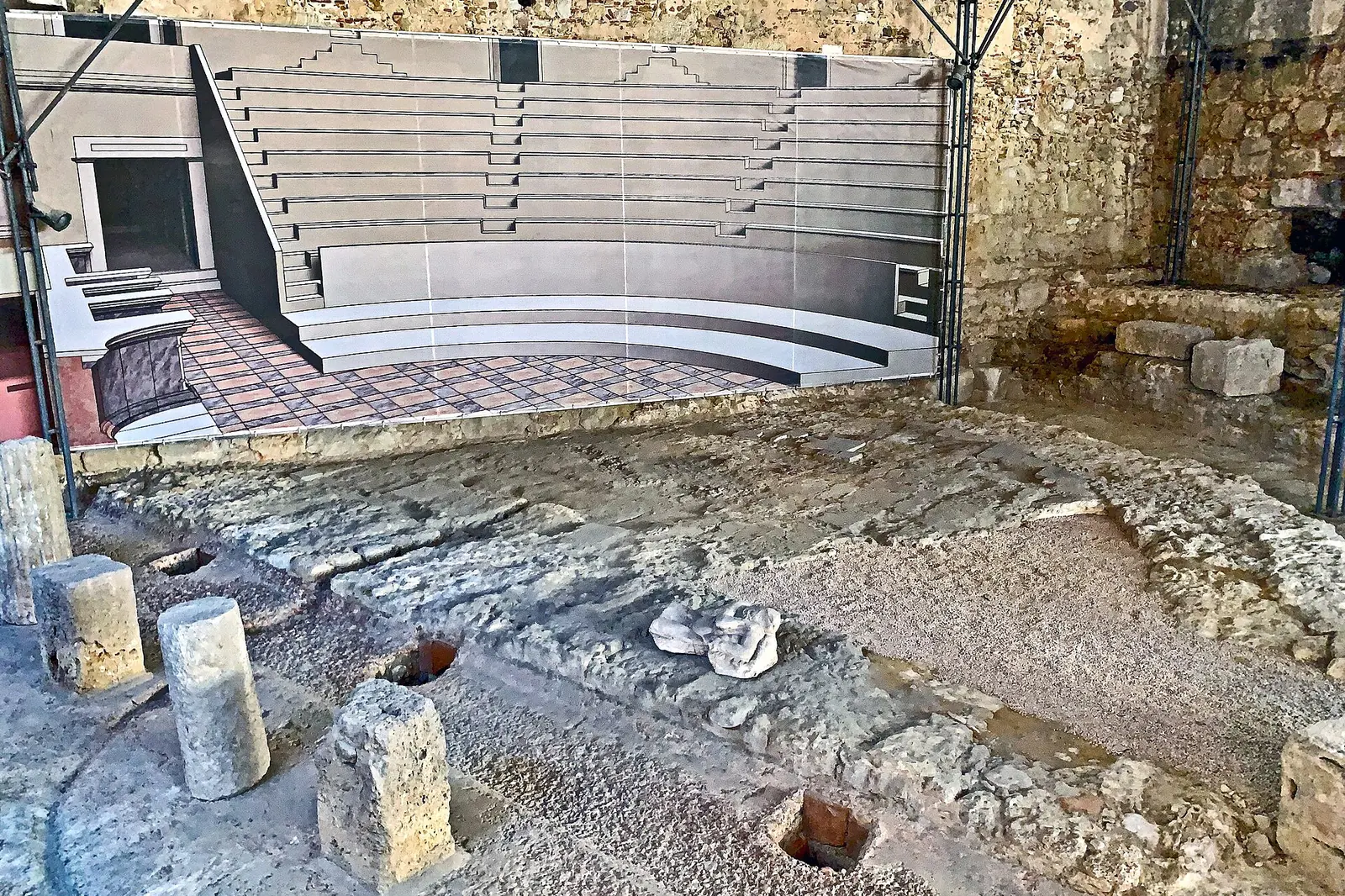 Excavated stone tiers and walls of the Roman theatre of Lisbon under a protective steel roof, with a model of the stage in the background